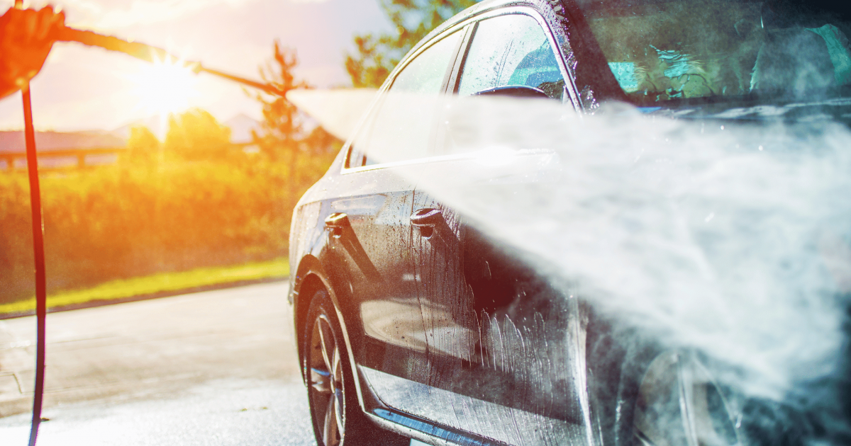 A black car being washed with a high-pressure hose under bright sunlight, illustrating the process of preparing your car for spring by removing winter salt and grime.